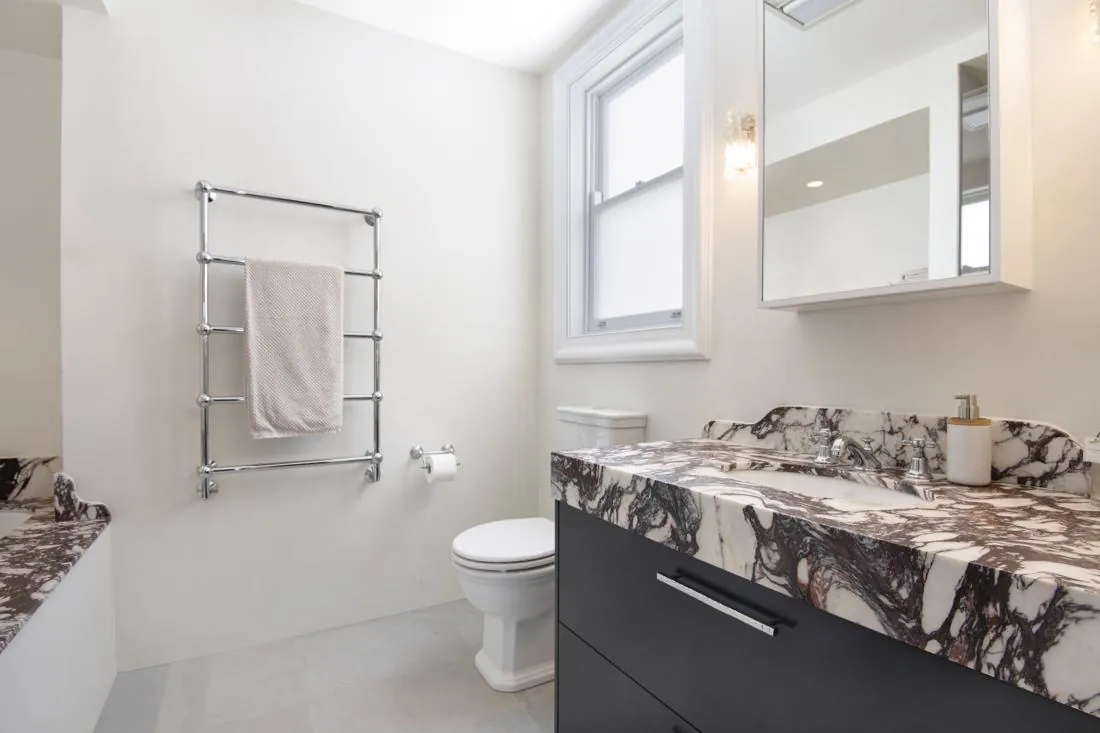 Bathroom with a marble countertop vanity, towel warmer, and toilet under a bright window; sleek, modern design.