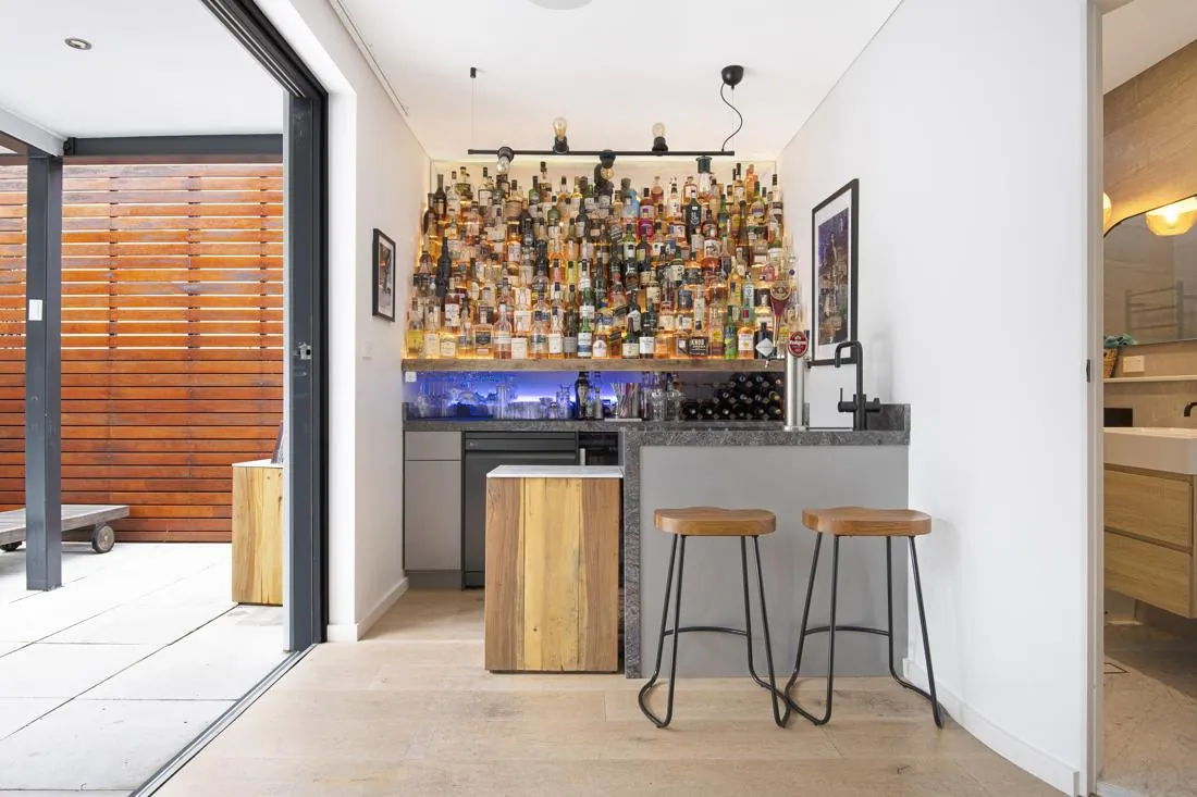 Modern home bar with a wall of liquor bottles, granite counter, and two wooden-stemmed stools by the bar