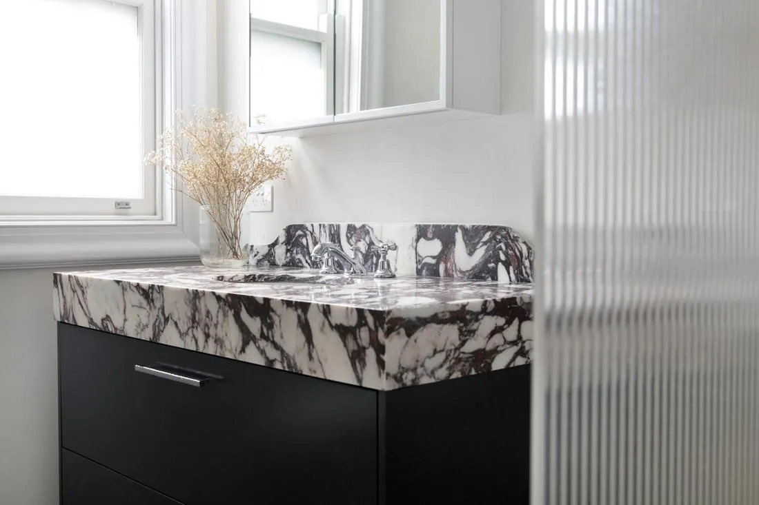 Bathroom vanity with black cabinets and a white marble countertop, chrome faucet, by a window with a vase of dried flowers nearby.