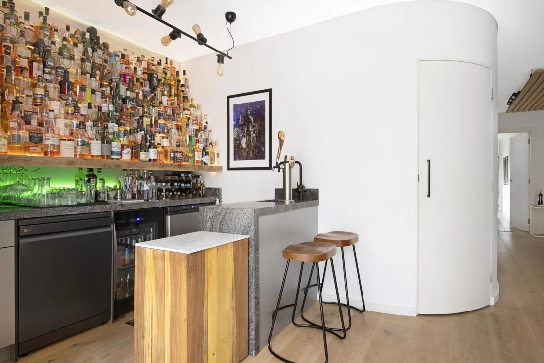 Bar area with a wall of liquor bottles, green under-shelf lighting, granite counter, and two wooden stools on metal legs.