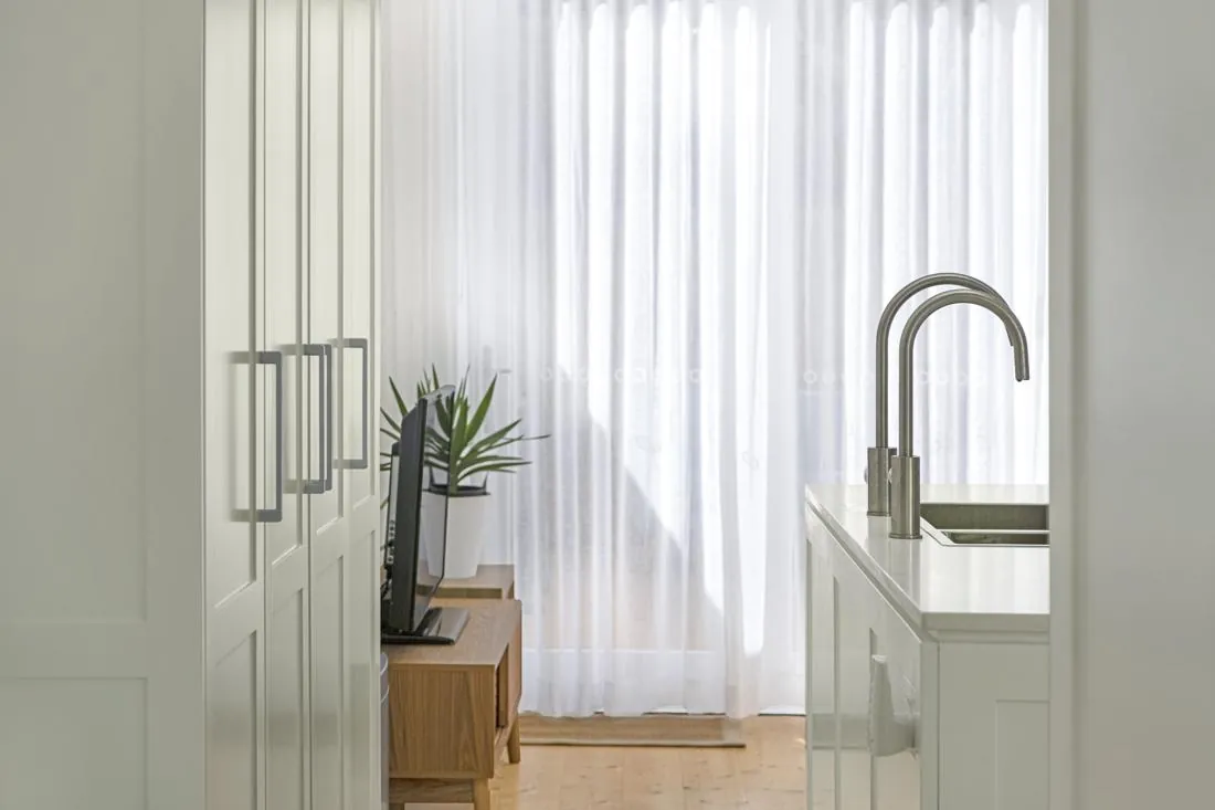 Bright modern kitchen with white cabinets, a stainless steel sink and curved faucet, and sheer white curtains behind.