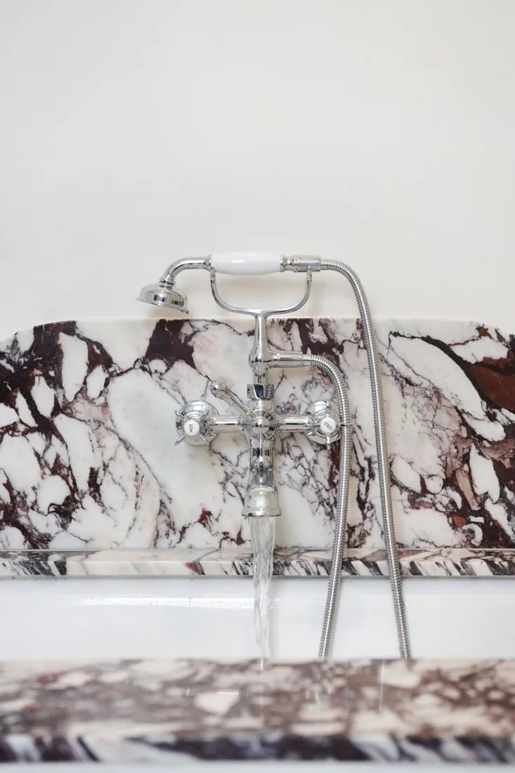 Chrome bathtub faucet with a handheld shower attachment mounted on a marble wall, water flowing into the tub.