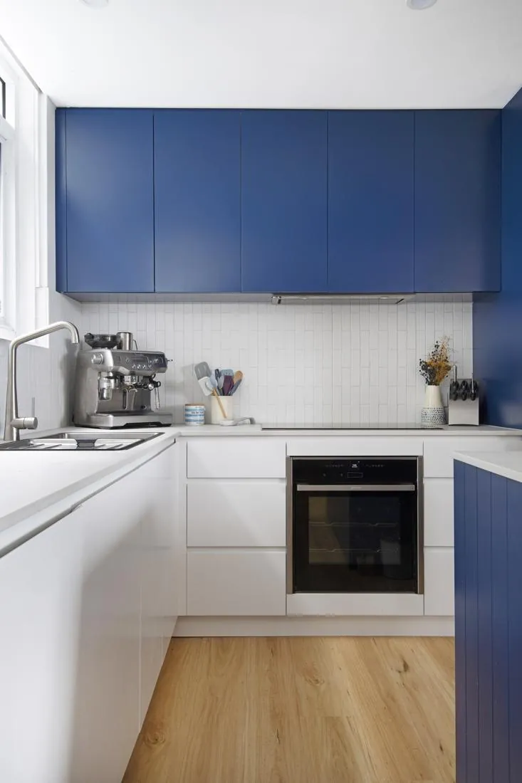 Modern L-shaped kitchen with white lower cabinets, blue upper cabinets, and a built-in oven under the counter. A silver espresso machine sits on the counter by the sink, with utensils and a vase in the background.